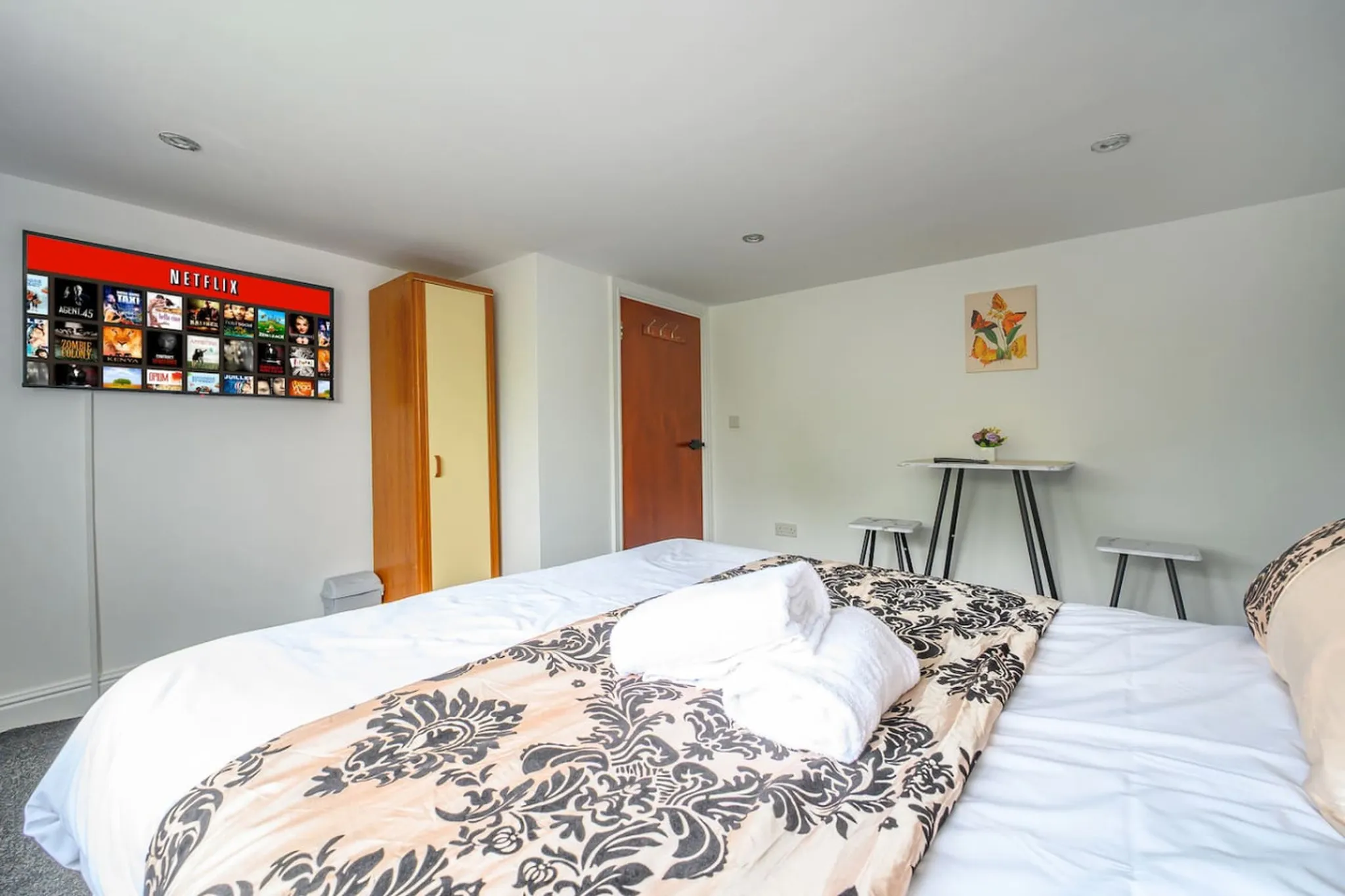 A neatly made bed in a bedroom with a TV, wardrobe, and a small table with stools.