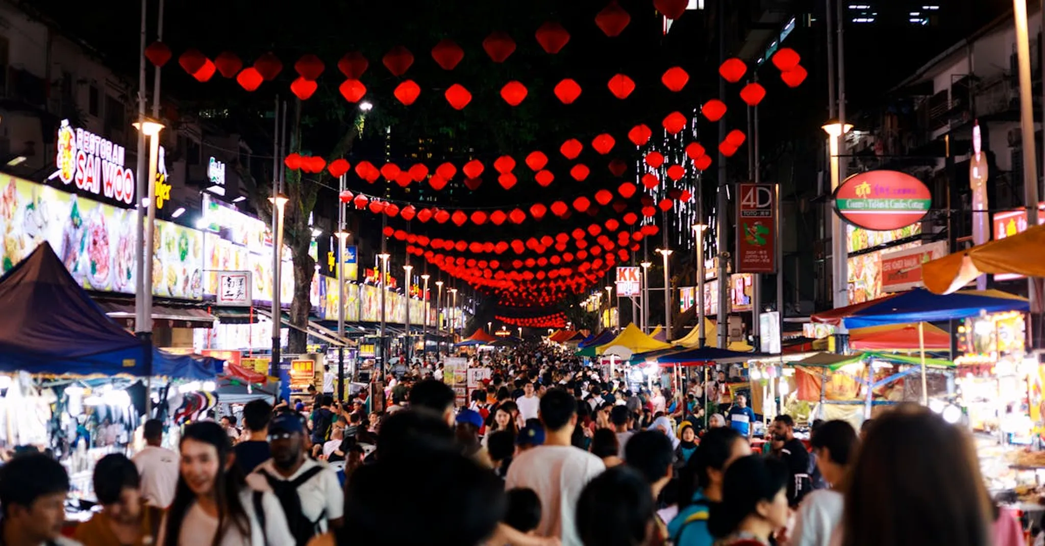 Bustling night market scene in Kuala Lumpur, featuring red lanterns and diverse crowd.