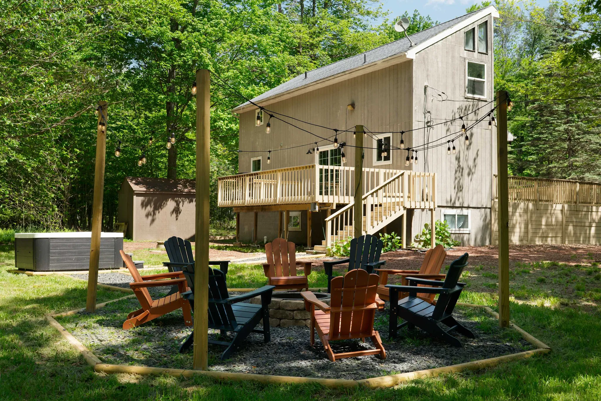 Backyard fire pit ringed with Adirondack chairs and string lights beside a two-story gray cabin with deck and hot tub in wooded surroundings