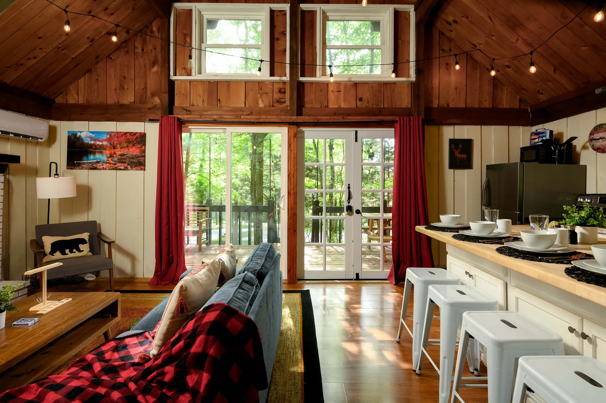 Open cabin interior with vaulted wood ceiling, red curtains framing glass doors to a forest view, sofa with buffalo-check throw, and