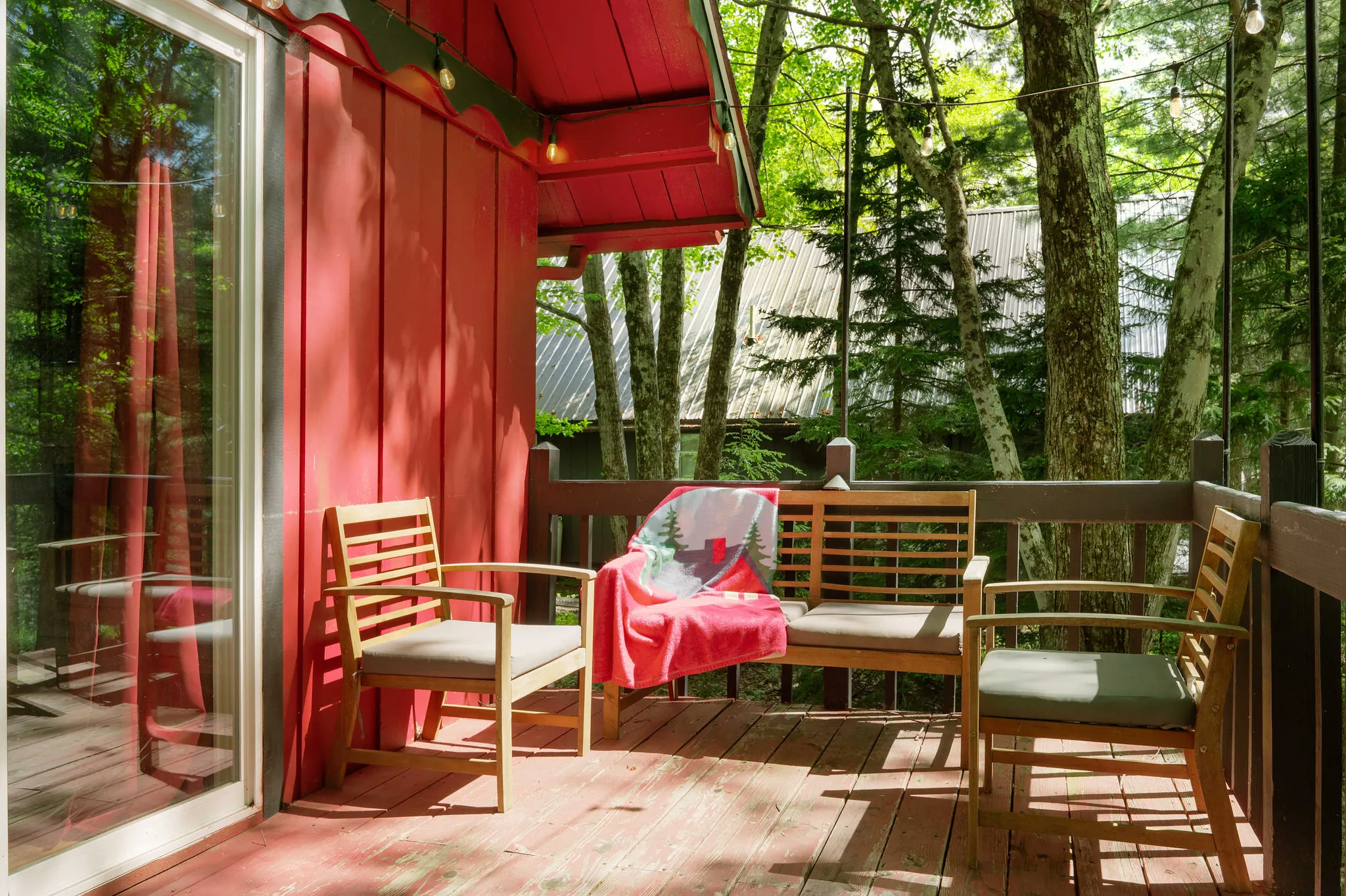 Red-painted cabin porch with wooden chairs, bench draped in pink blanket, string lights, and forest views through tall trees