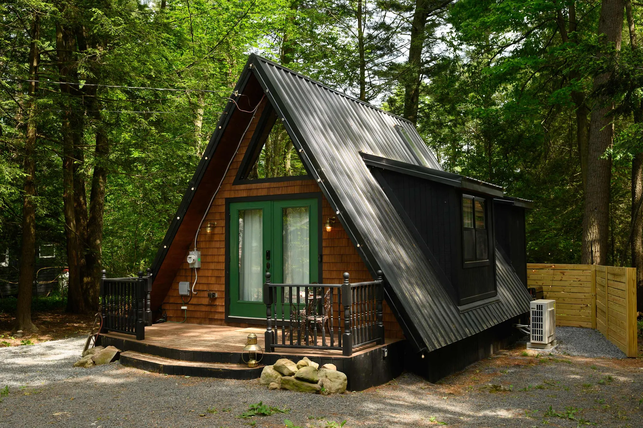 A-frame cabin with dark metal roof, green double doors, and wood deck nestled among tall forest trees