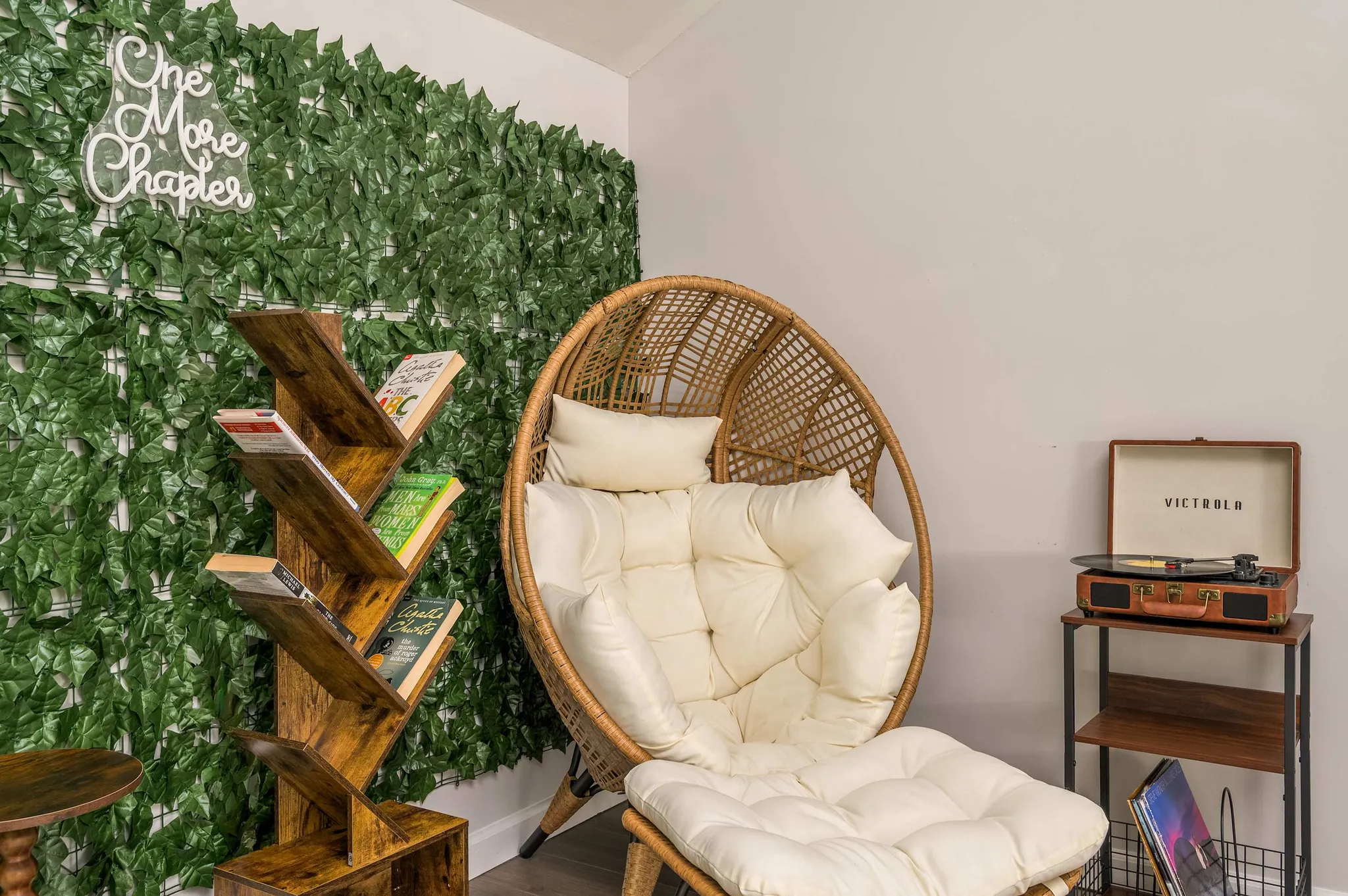 Cozy reading nook with rattan egg chair, tree-shaped bookshelf, faux ivy wall, neon "One More Chapter" sign, and vintage record player