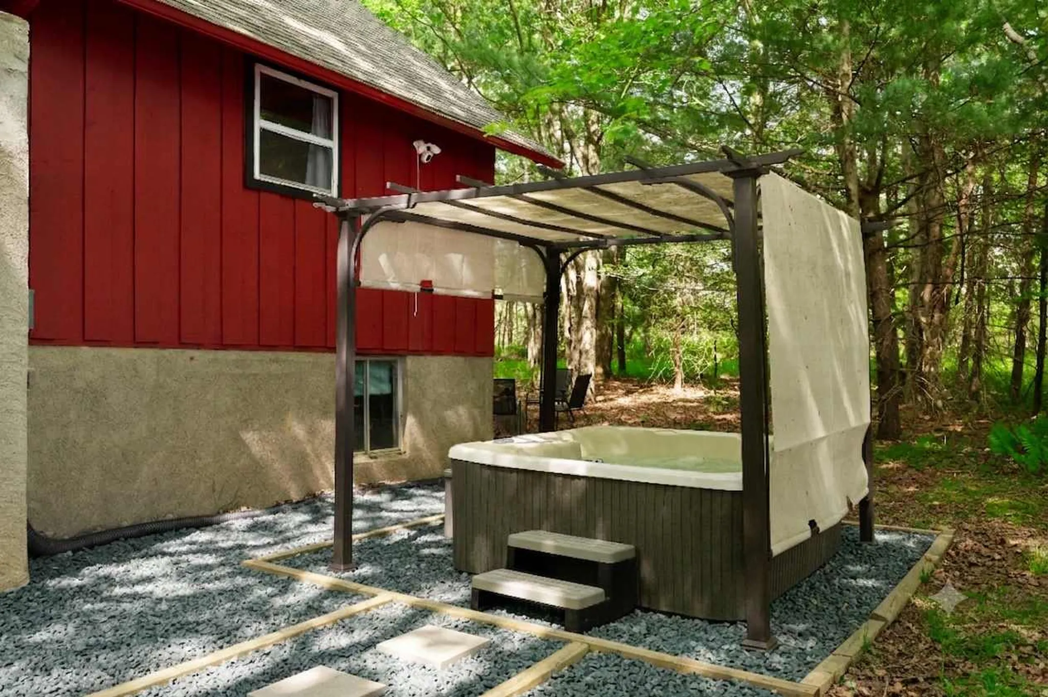 Outdoor hot tub with steps under a white pergola beside a red barn-style cabin, set on gravel patio in wooded surroundings
