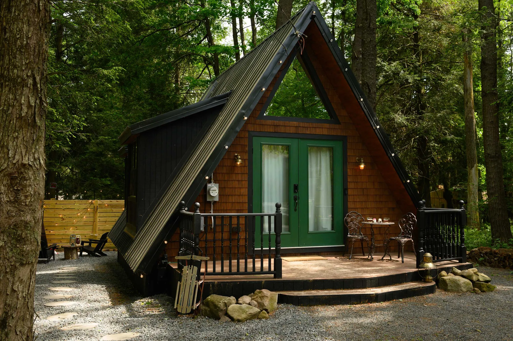 A-frame cabin with green double doors, string lights, and a small deck with bistro chairs nestled among tall forest trees