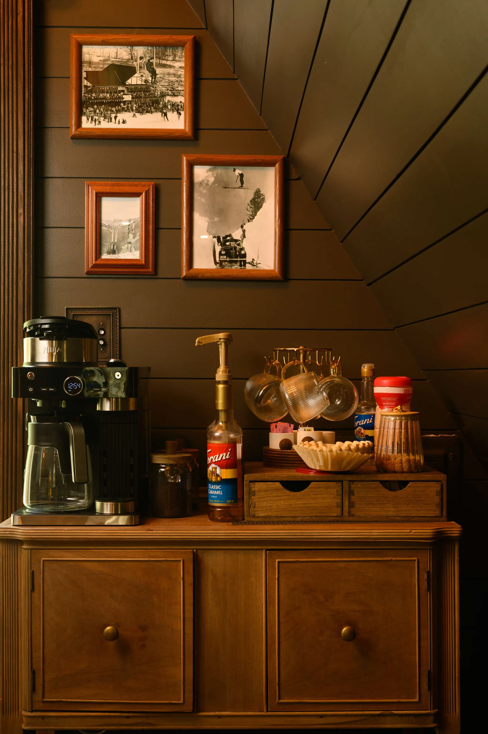 Cozy A-frame coffee bar on wooden cabinet with espresso machine, syrup bottle, hanging mugs, and framed vintage ski photos on dark paneled