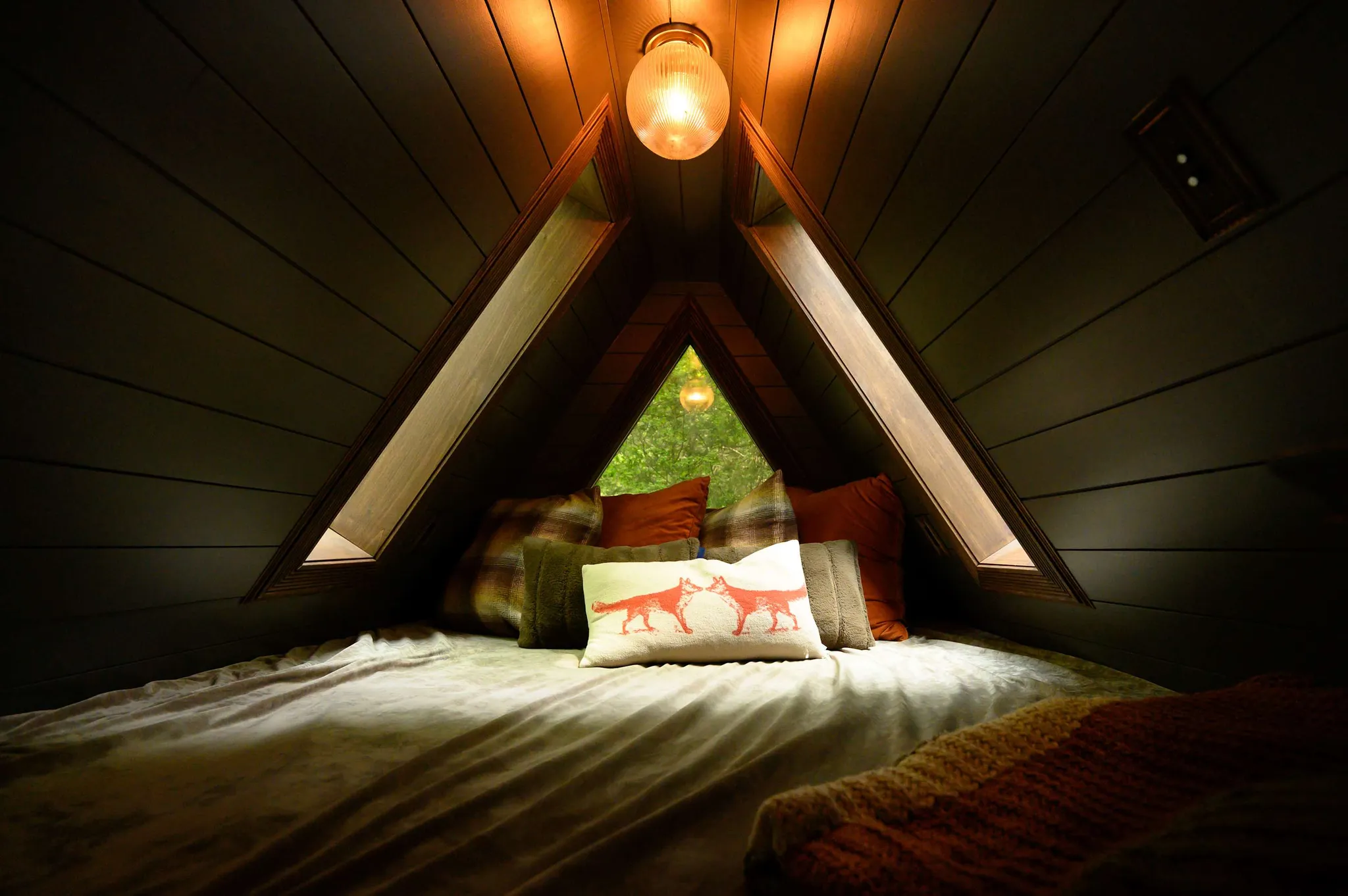 Cozy A-frame cabin loft bed with pendant light, plaid pillows, and fox-print cushion framed by triangular window
