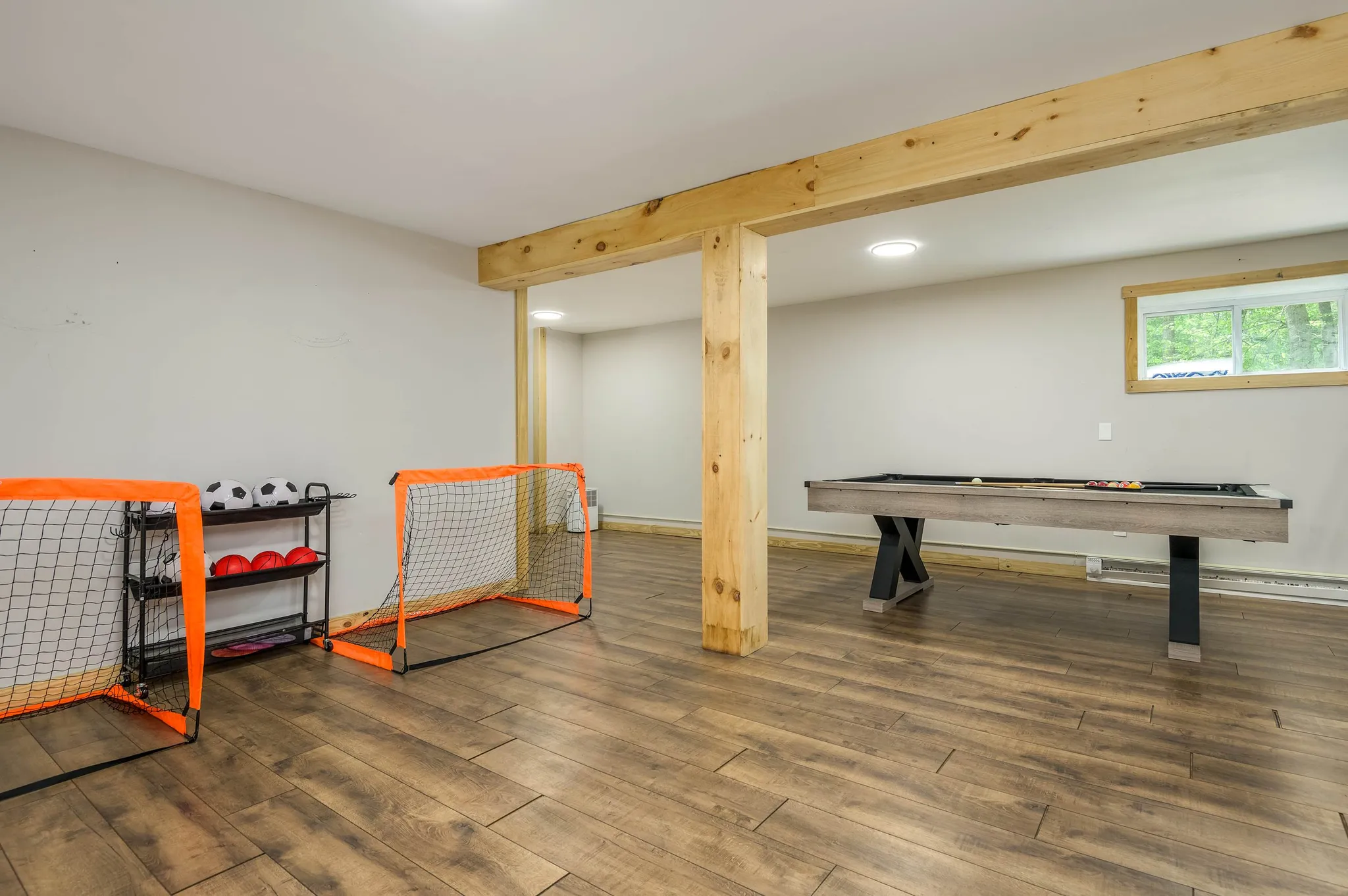 Spacious basement game room with mini soccer goals, balls, and a pool table on wide-plank wood flooring under exposed beams