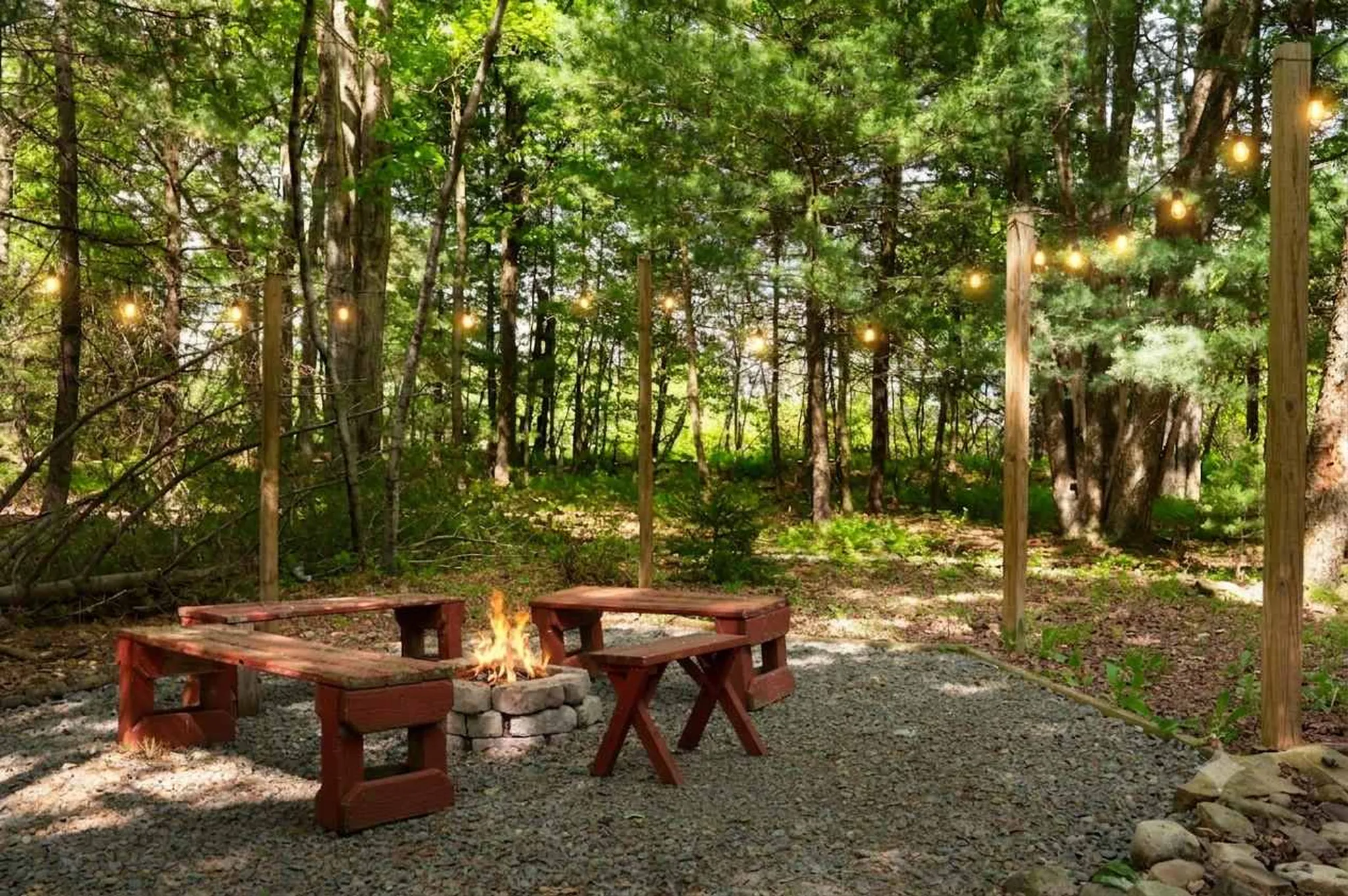 Wooded backyard fire pit area with wooden benches, gravel ground, and string lights hung between tall posts among trees