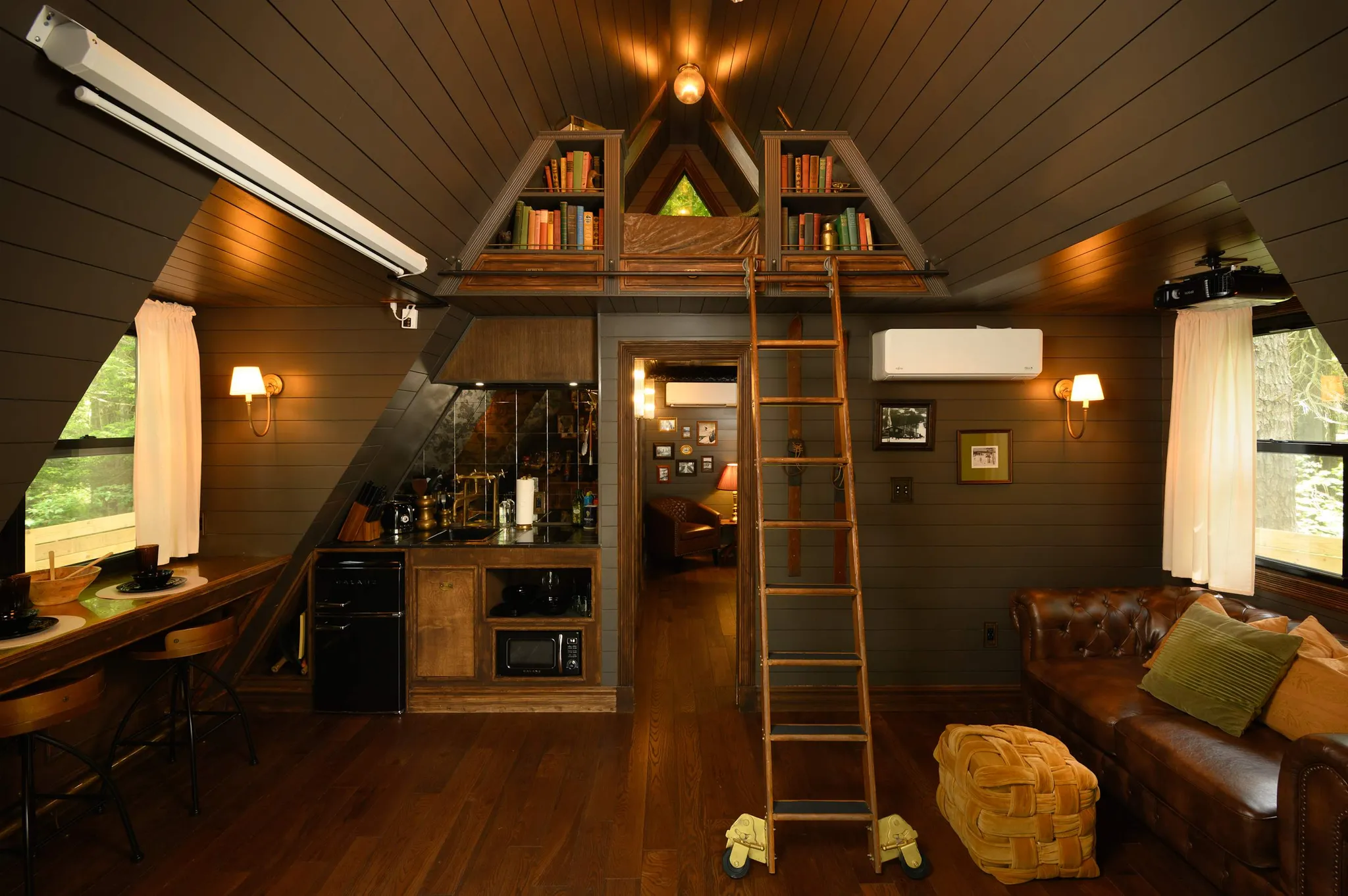 A-frame cabin interior with wooden walls, loft ladder to triangular bookshelves, kitchenette, leather sofa, and bar stools at counter