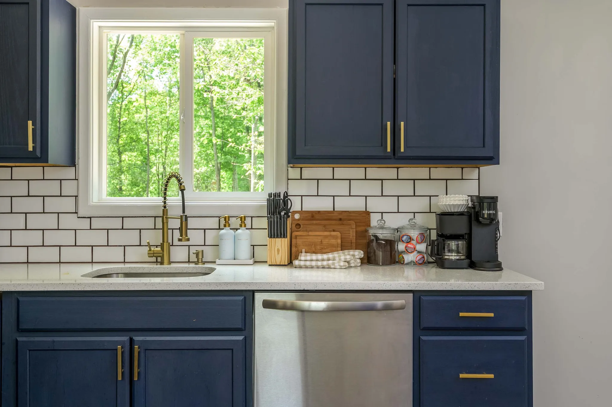 Navy blue kitchen with white subway tile backsplash, farmhouse sink under a window, stainless dishwasher, and coffee maker on quartz counter