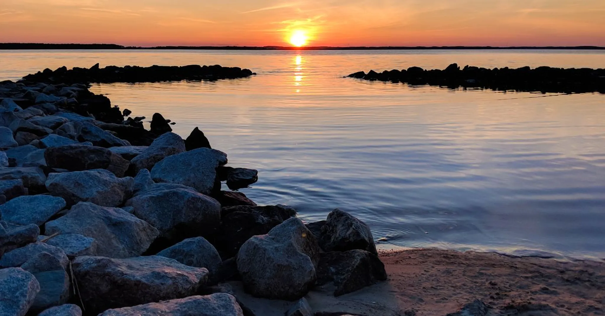 Sunset over calm bay water with rocky shoreline jetty in the foreground and orange sky reflecting on the surface