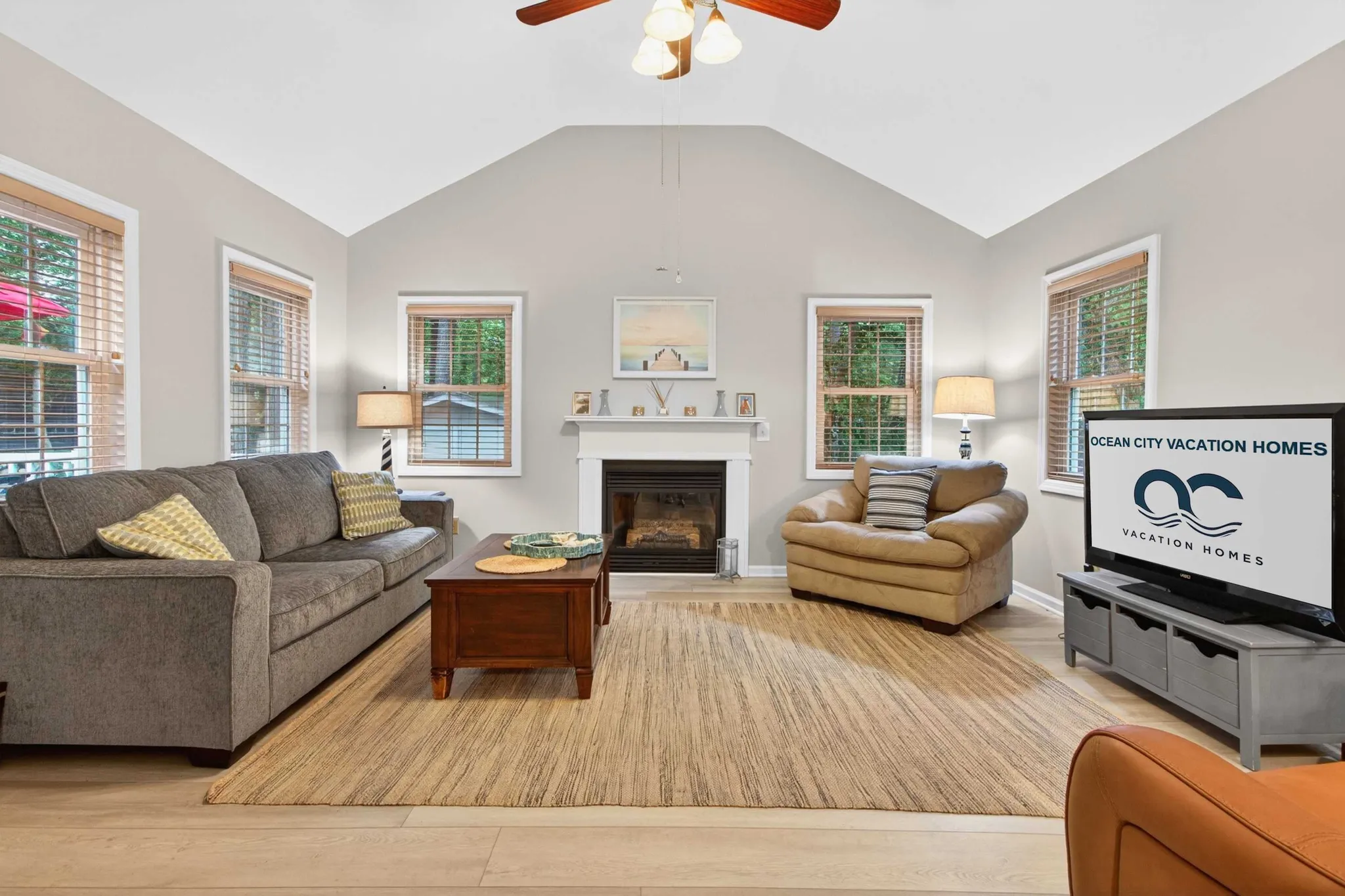 Vaulted-ceiling living room with gray sofa, armchair, fireplace, ceiling fan, and TV displaying Ocean City Vacation Homes logo