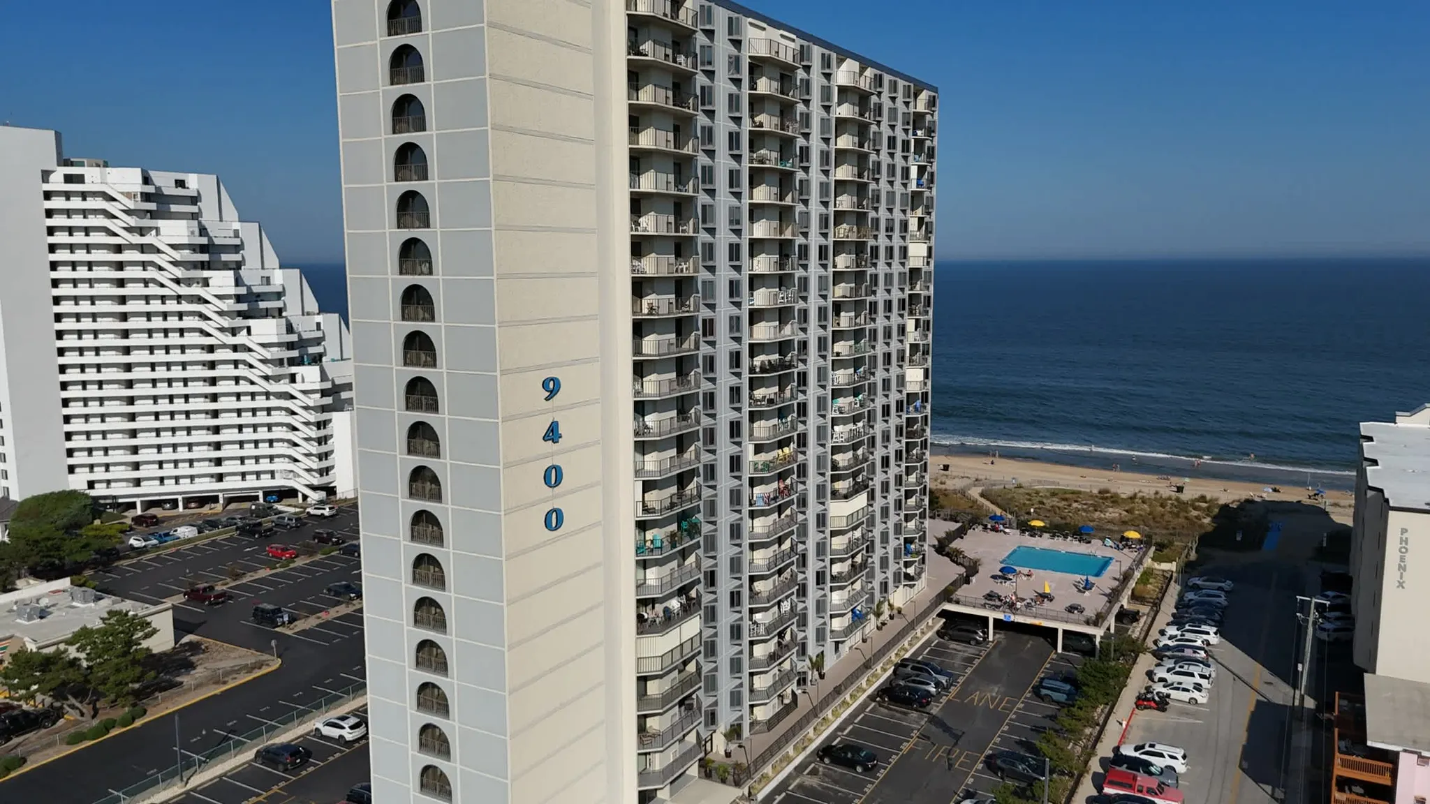 Aerial view of a tall beachfront condominium tower marked 9400 with balconies, oceanfront pool, and parking lots beside the shoreline