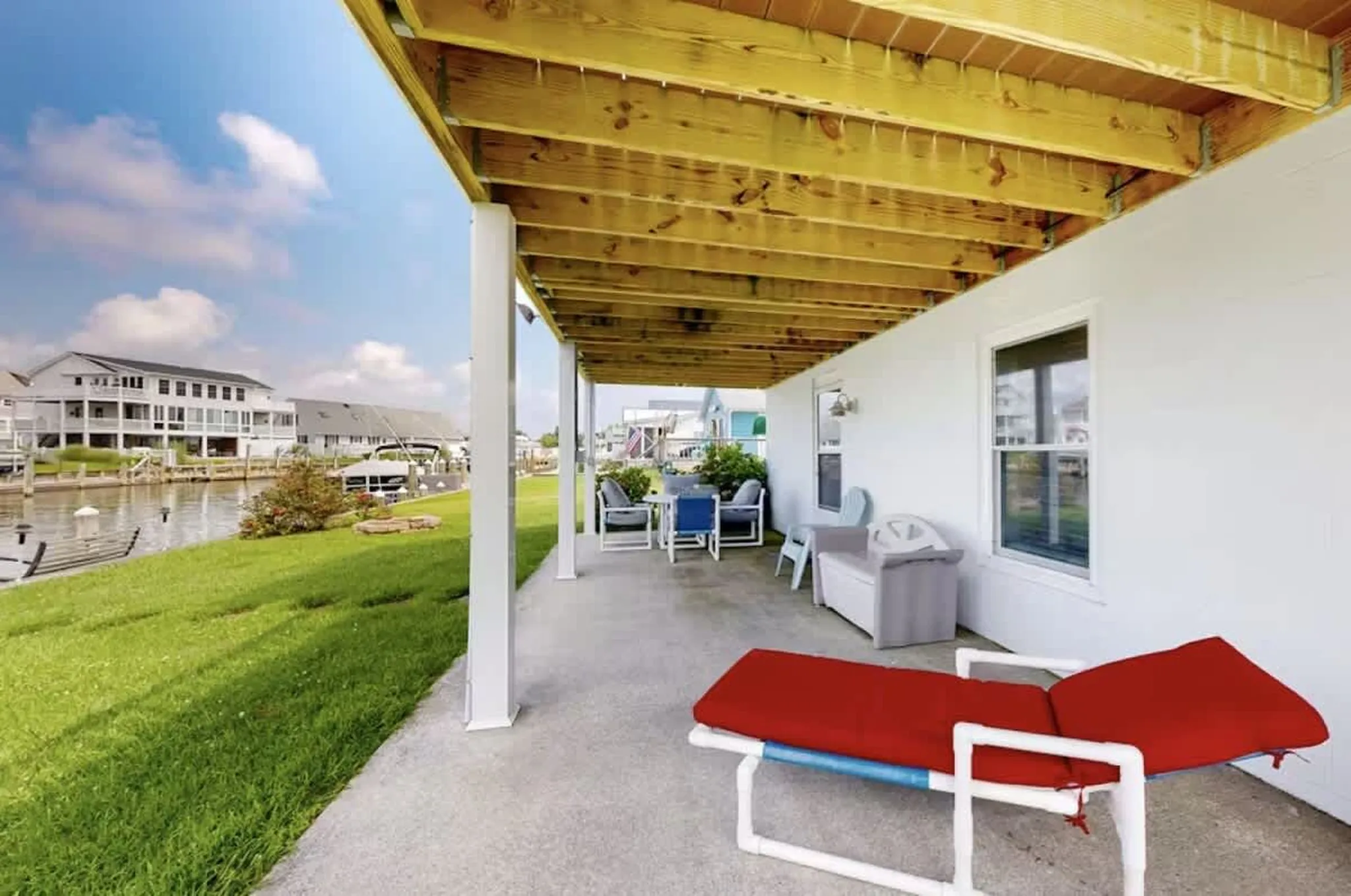 Covered waterfront patio with red lounge chair, outdoor seating, and canal view of neighboring homes and docked boats