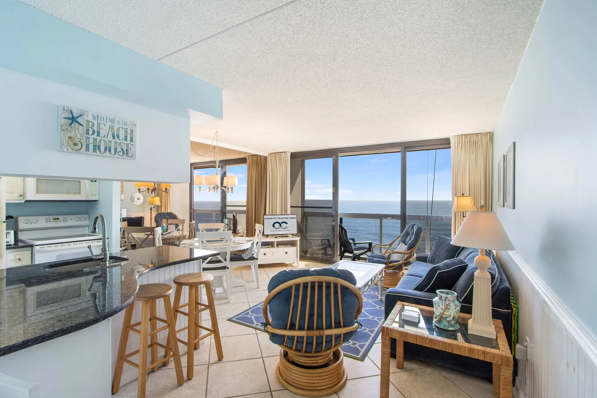 Open-plan beachfront condo living area with rattan chairs, blue sofa, kitchen bar stools, and floor-to-ceiling ocean view windows