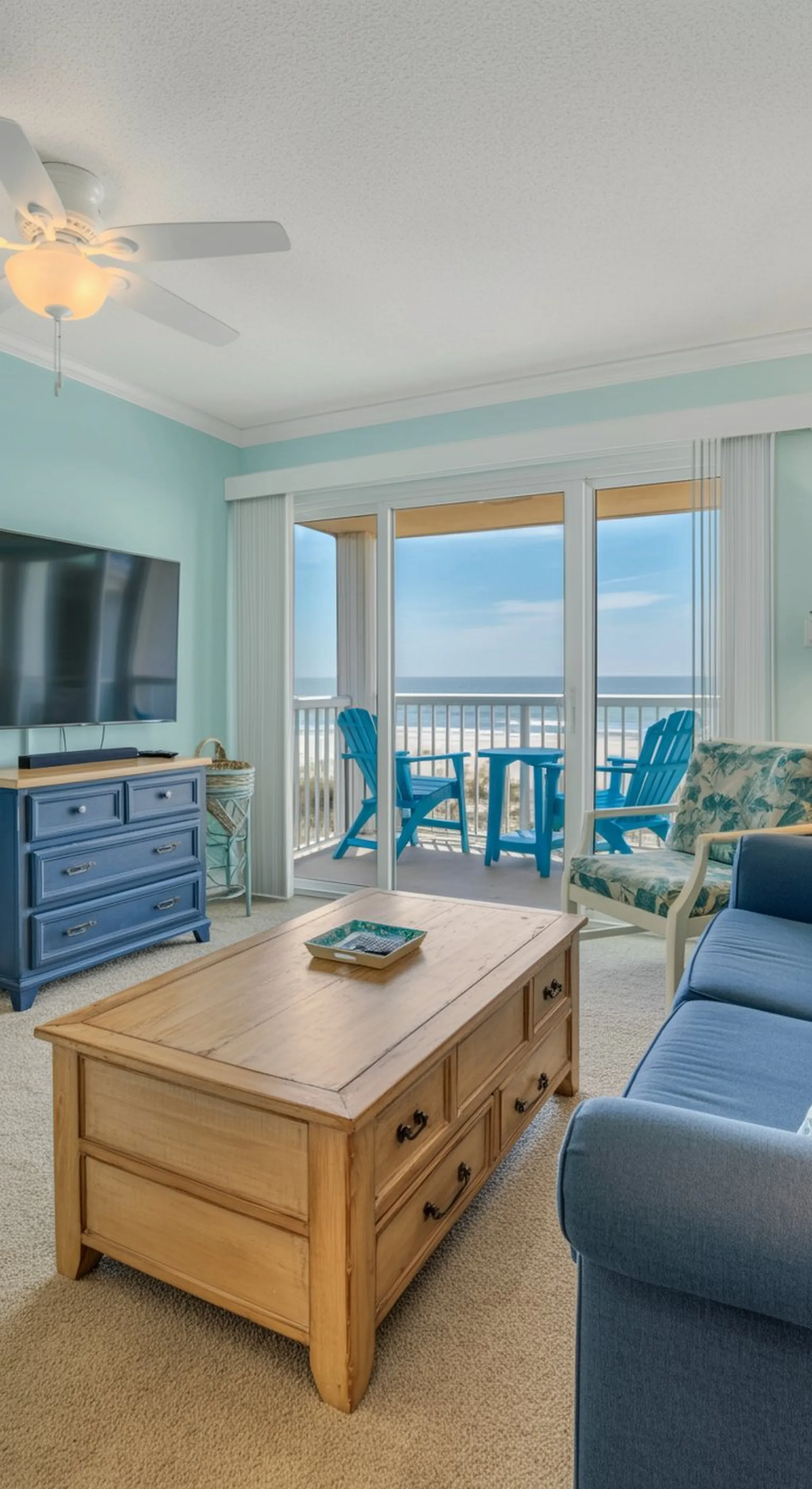 Beachfront living room with ceiling fan, blue dresser, TV, wooden coffee table, and balcony with Adirondack chairs overlooking the ocean