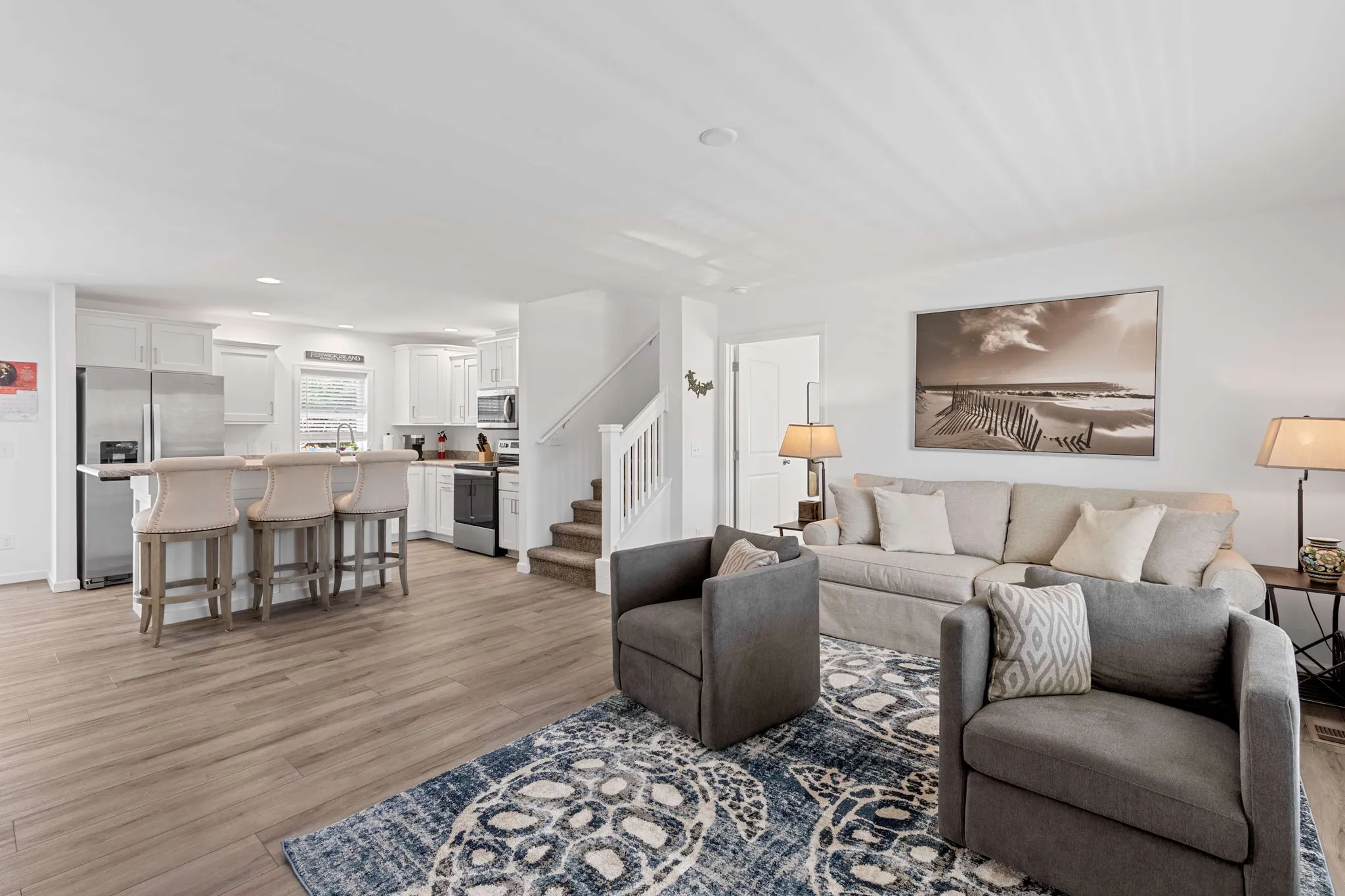 Open-concept living room with gray sectional, accent chairs, patterned rug, and view into white kitchen with island seating and staircase