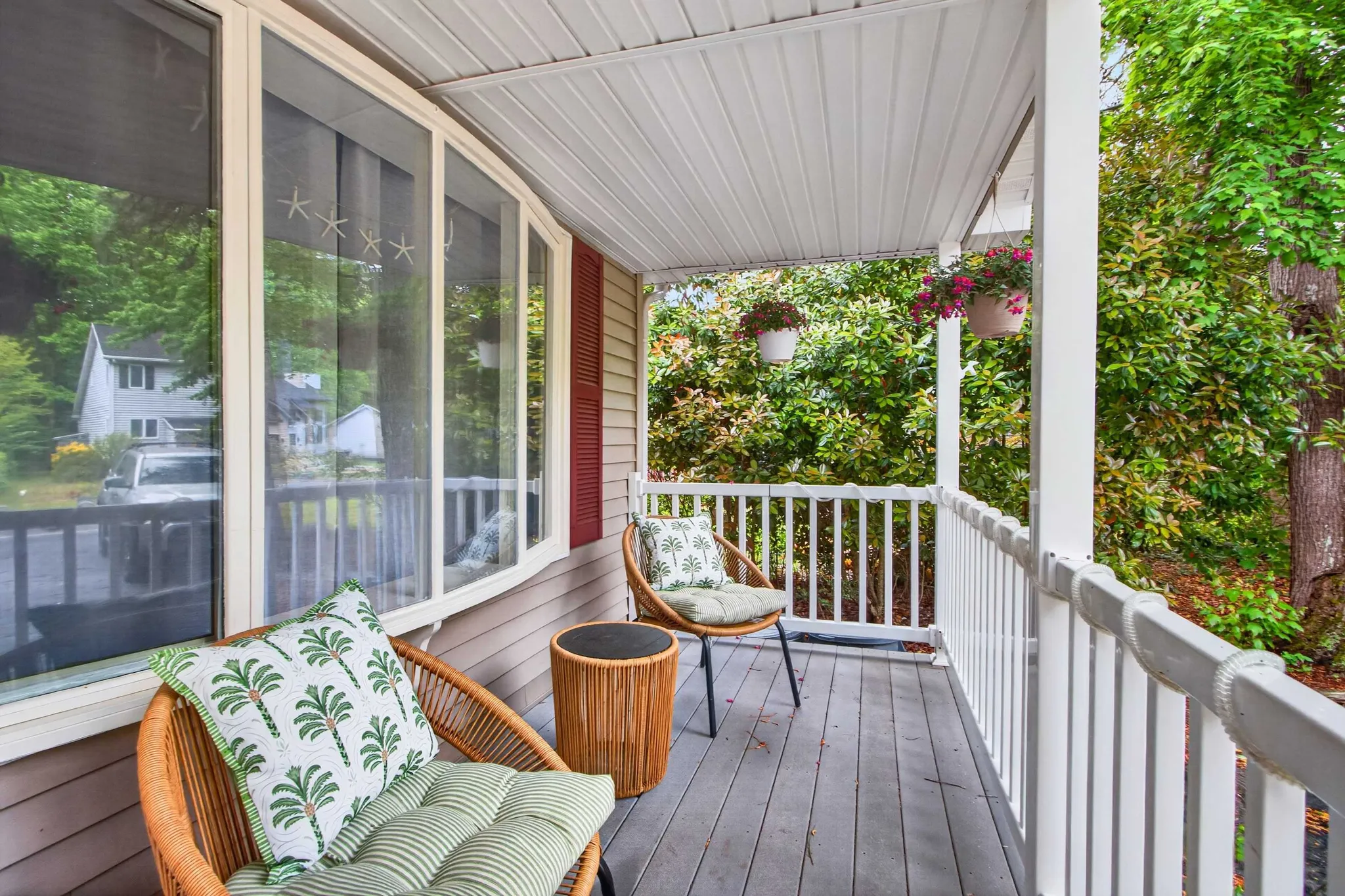 Covered porch with wicker seating, palm-print pillows, hanging flower baskets, and white railing overlooking a leafy neighborhood