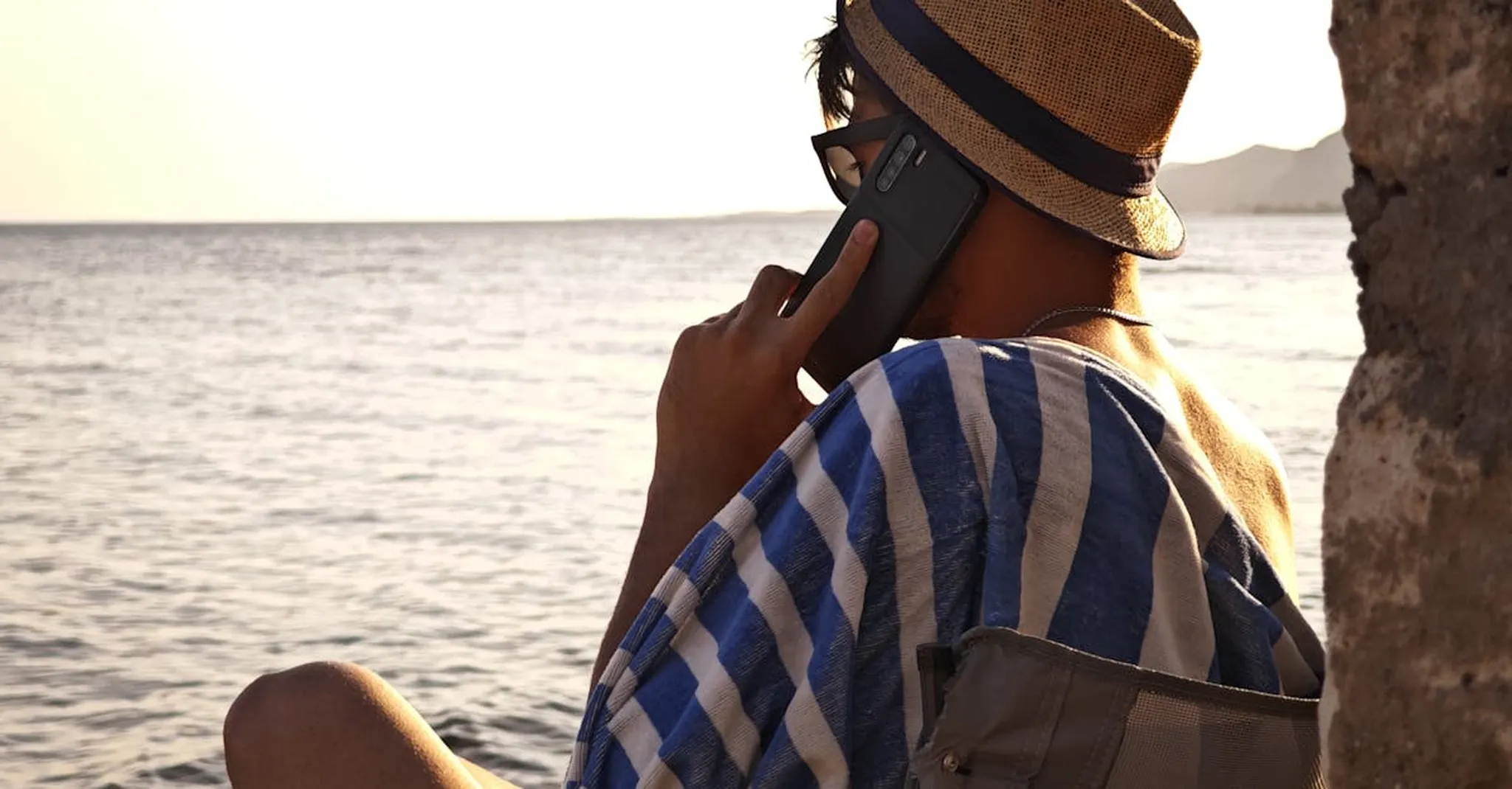 A man wearing a straw hat talks on a smartphone by the sea. Perfect summer vacation vibe.