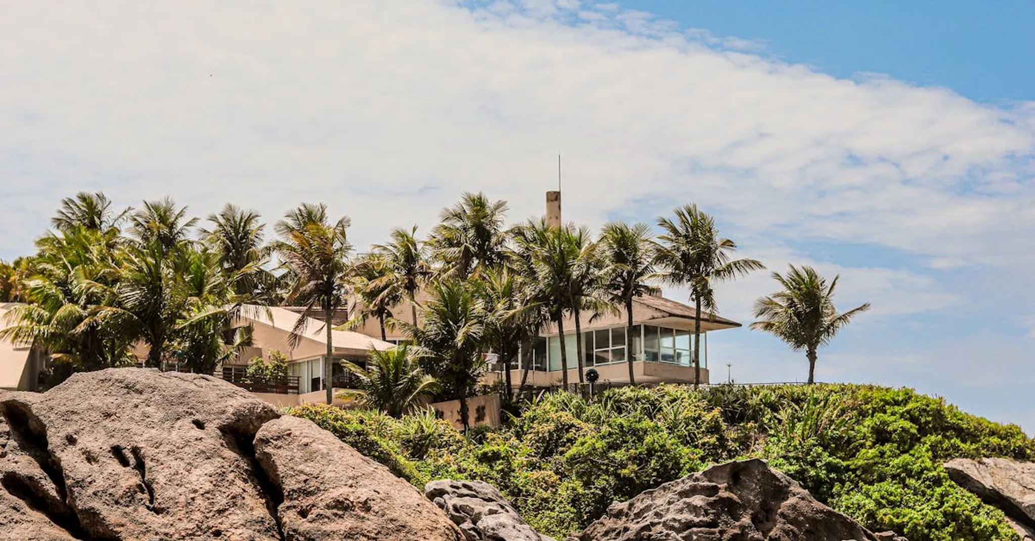 A stunning tropical villa surrounded by palm trees on a rocky coastline under a bright sky.