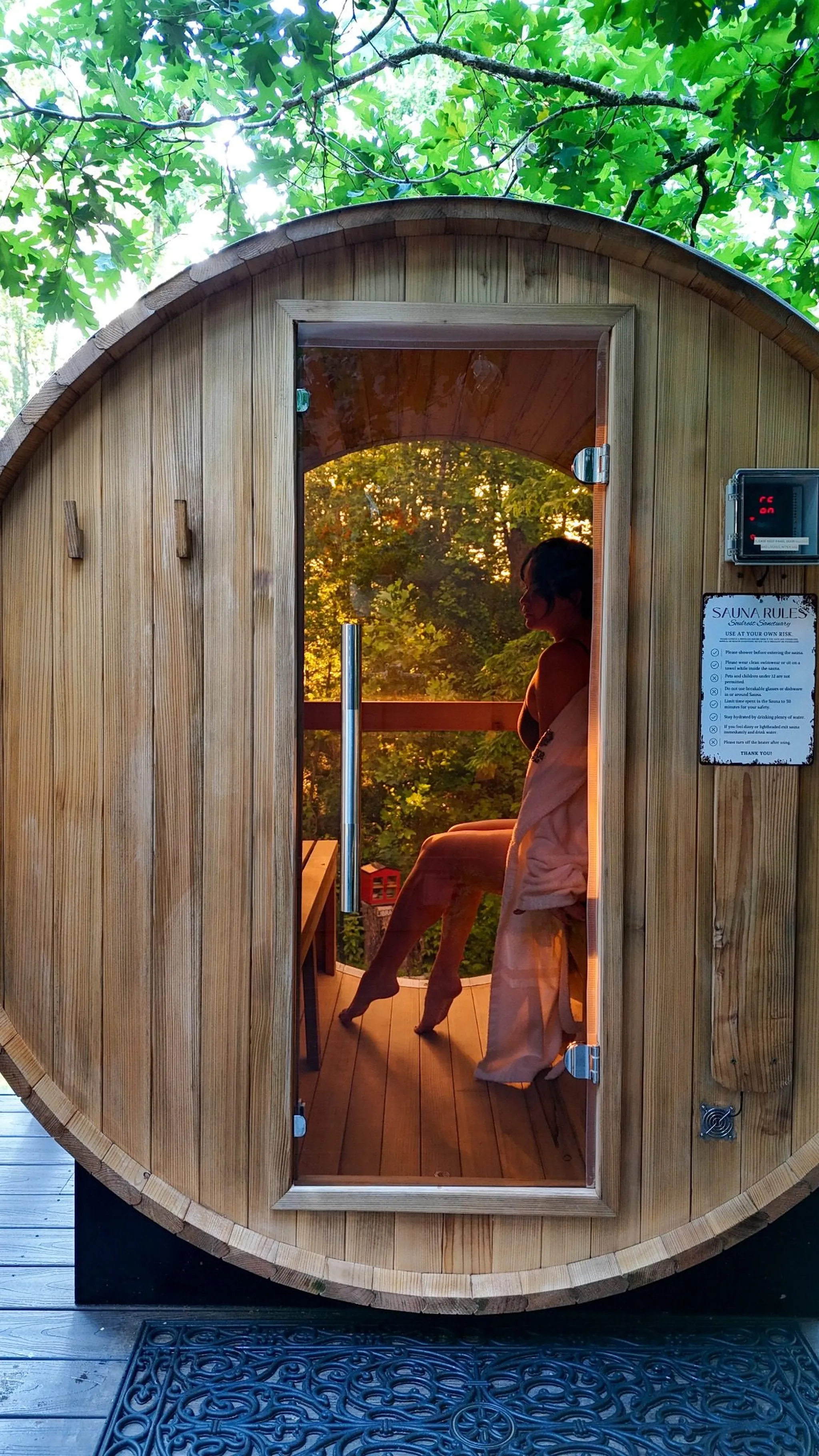 Wooden barrel sauna and outdoor shower on a deck surrounded by trees.