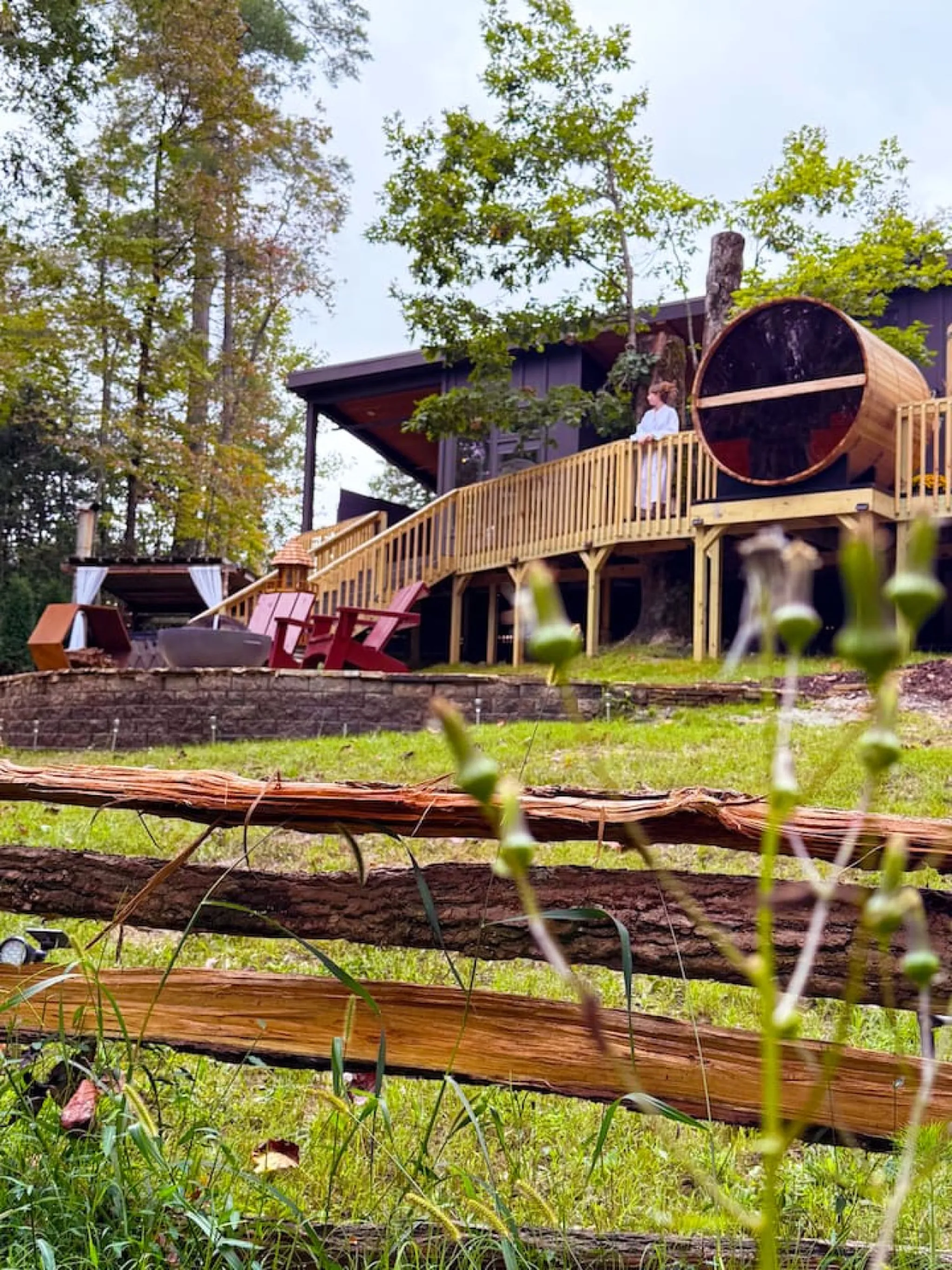 Modern treehouse with a barrel sauna and a wooden deck, surrounded by trees.