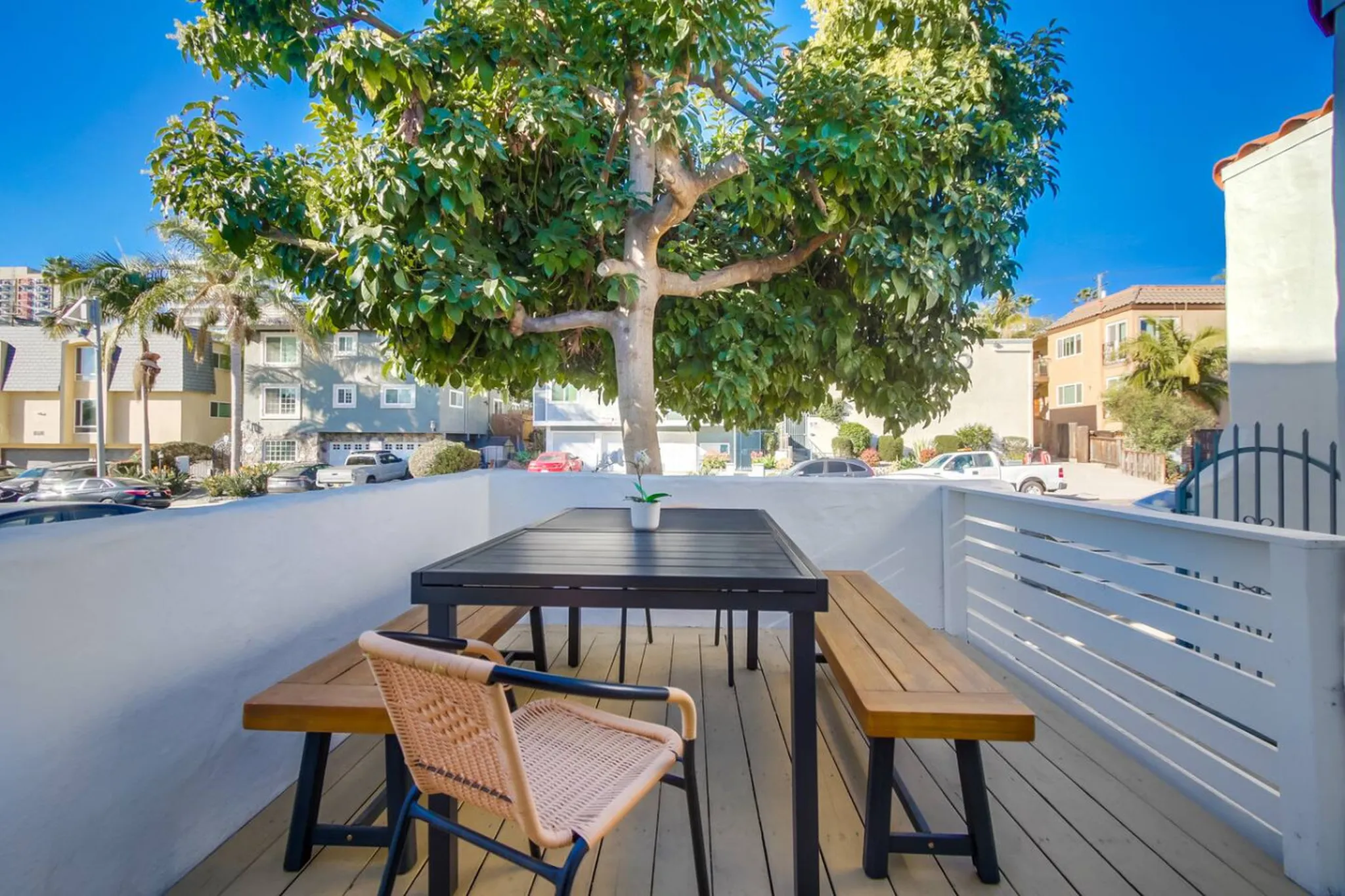 Rooftop deck with wooden dining table, benches, woven chair, and potted plant shaded by a large tree overlooking a neighborhood street