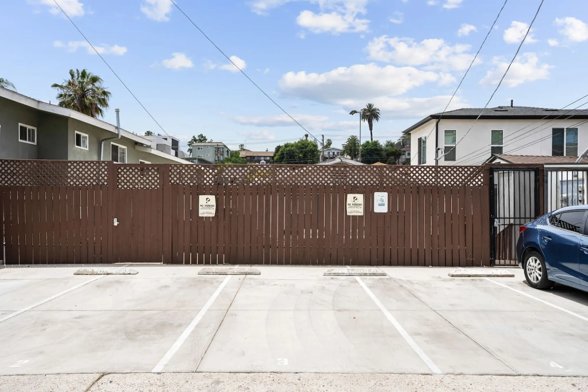 Private parking area with numbered spaces behind a tall wooden fence, palm trees and houses visible beyond